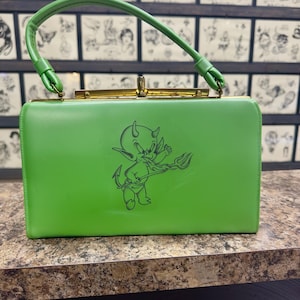 May include: Bright green handbag with a matching handle and gold-colored clasp. The bag features a cartoon devil graphic on the front. The bag is rectangular and appears to be made of a smooth material.