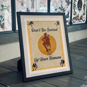 May include: A framed print with a yellow background and black text that reads "Don't Be Scared Of Your Demons". A red demon with horns and a tail is sitting on a stool playing a banjo. There are black silhouettes of demons in the corners of the print.