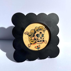 May include: A framed tattoo-style image featuring a skull with crossed bones and a butterfly. The artwork is set within a black, scalloped-edge frame. The skull has a vintage aesthetic with tan, yellow, and black colors.