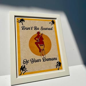 May include: Framed art print with a white border. The artwork features a red devil playing a banjo on a stool, set against a yellow circle and a tan background. The text "Don't Be Scared Of Your Demons" is displayed.