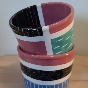 Könnte beinhalten: Zwei Keramikbecher, die übereinander gestapelt sind. Der obere Becher ist rosa, weiß, schwarz und grün mit geometrischen Mustern. Der untere Becher ist blau, weiß und schwarz mit Streifen und einem Blumenmuster.