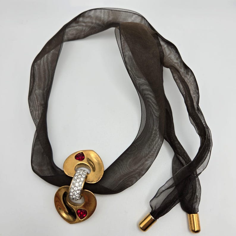 May include: A gold-tone necklace with a double heart pendant featuring clear rhinestones and red stones. The necklace is on a black ribbon.