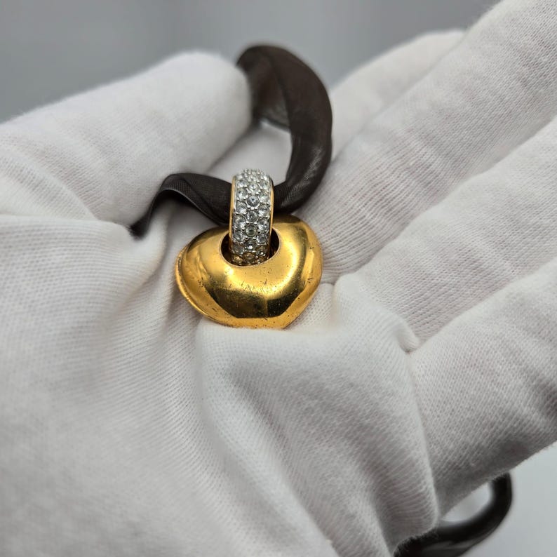 May include: A gold heart-shaped pendant with a clear crystal-encrusted band. The pendant is attached to a brown ribbon.