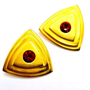May include: Two gold-toned, triangular earrings with a layered design. Each earring features a central, round, red gemstone. The earrings are set against a white background, highlighting their metallic sheen and gemstone sparkle.