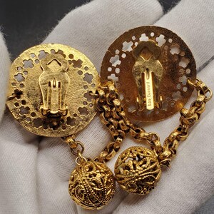 May include: Gold tone clip-on earrings with a filigree design. The earrings feature two round, openwork discs connected by a chain with two dangling filigree balls. The back of one disc has the inscription "Les Bernard".