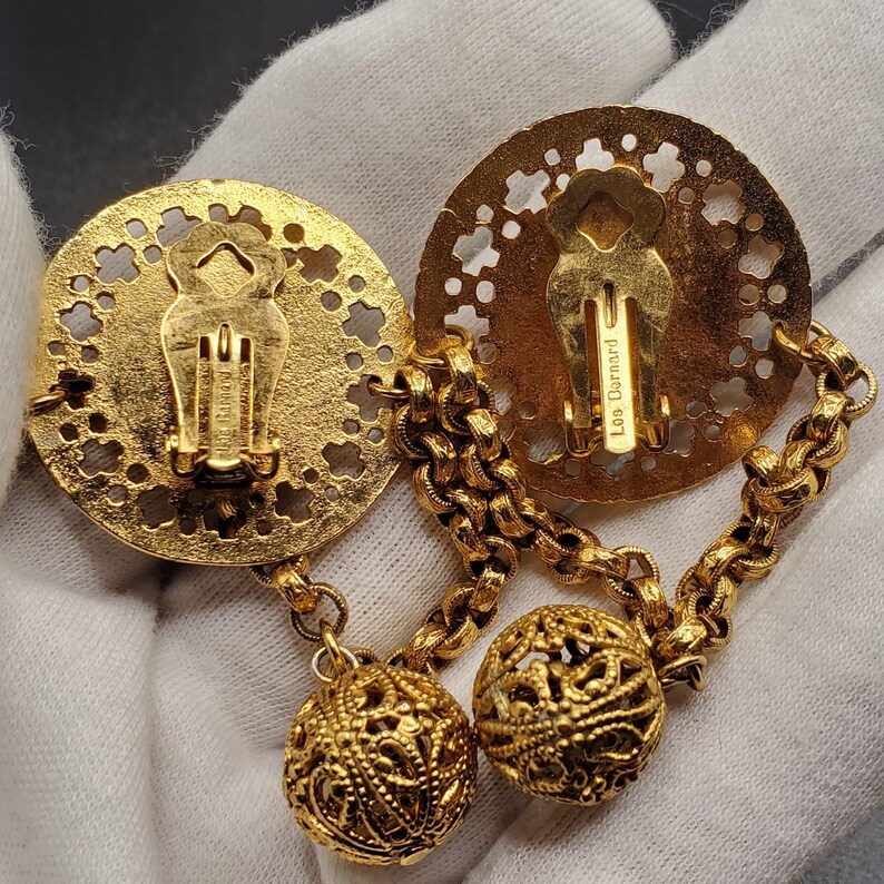 May include: Gold tone clip-on earrings with a filigree design. The earrings feature two round, openwork discs connected by a chain with two dangling filigree balls. The back of one disc has the inscription "Les Bernard".