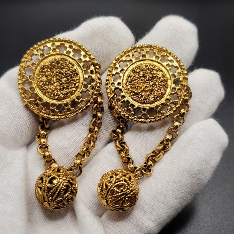 May include: A pair of gold-tone earrings with a circular design and a dangling ball. The earrings are made of metal and have a filigree pattern.