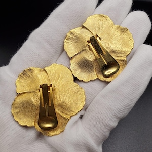 May include: A pair of gold-tone flower-shaped clip-on earrings. The earrings are made of metal and have a textured surface.