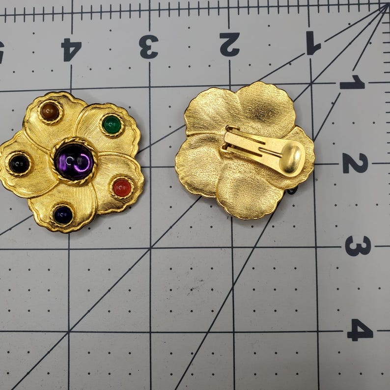 May include: A pair of gold tone clip-on earrings with a flower design. The earrings have five colorful stones set in the flower petals. The back of the earrings have a simple clip closure.