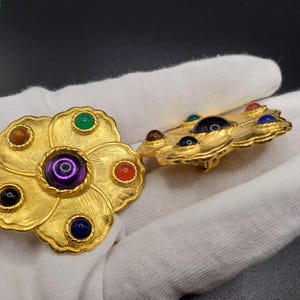May include: A pair of gold tone flower-shaped brooches with colorful stones. The brooches have a raised, textured design and feature a variety of colors, including purple, green, blue, red, and brown.