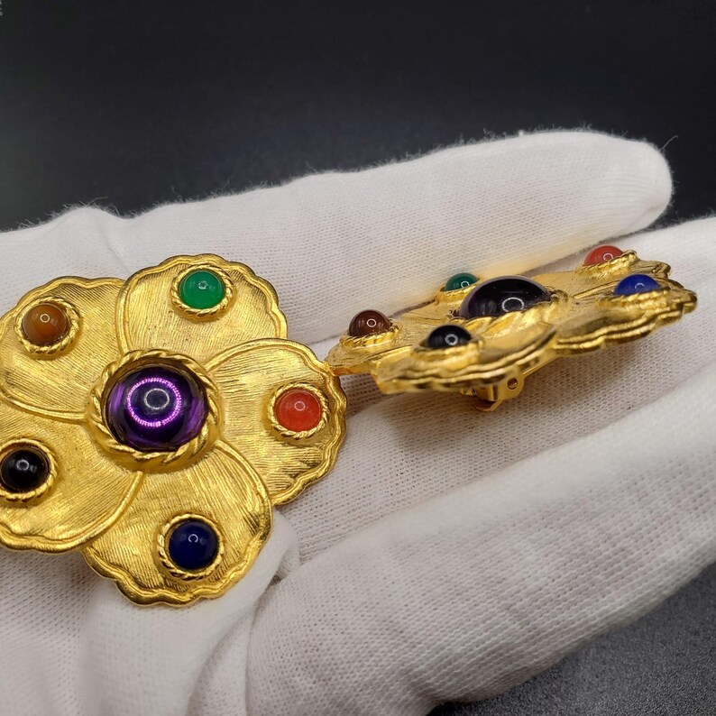 May include: A pair of gold tone flower-shaped brooches with colorful stones. The brooches have a raised, textured design and feature a variety of colors, including purple, green, blue, red, and brown.
