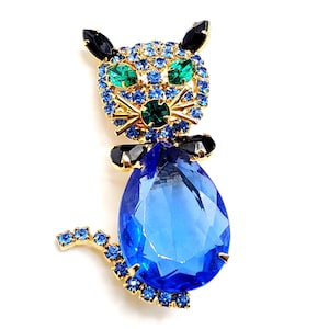 May include: A gold-tone brooch in the shape of a cat with a large blue teardrop-shaped rhinestone. The cat has black ears, green eyes, and blue rhinestones for its body and tail.