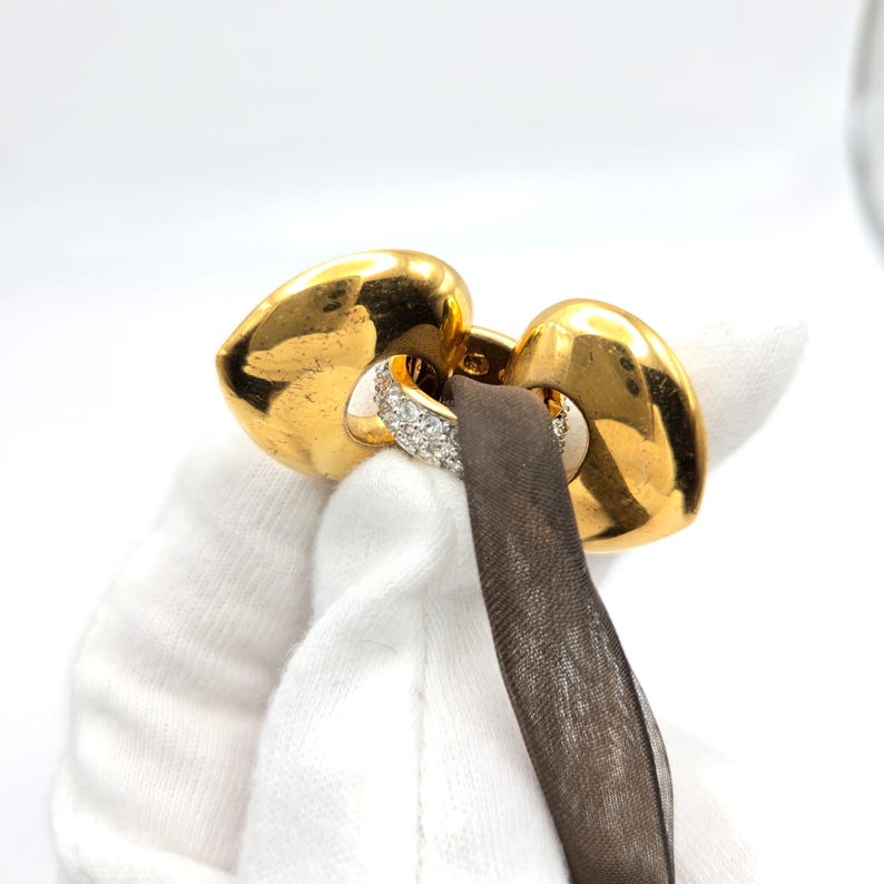 May include: A gold heart-shaped pendant with a diamond-encrusted band. The pendant is attached to a black ribbon.