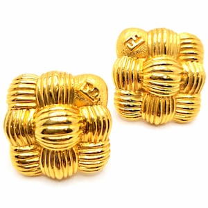 May include: A pair of gold-toned square earrings. Each earring features a woven design with a central, textured sphere and a smaller, embossed logo. The earrings have a bright, polished finish, reflecting light.