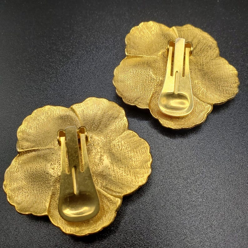 May include: A pair of gold-tone flower-shaped clip-on earrings. The earrings have a textured surface and a simple, classic design.