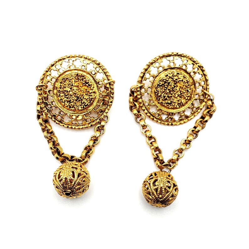 May include: A pair of gold-tone earrings with a filigree design. Each earring features a round, openwork pendant with a smaller, textured disc in the center. The pendants are attached to a chain that ends in a round, openwork ball.