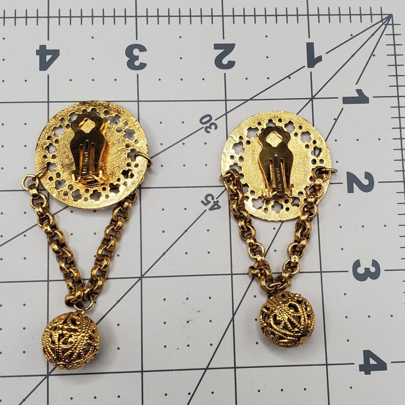 May include: A pair of gold tone earrings with a round, openwork design and a dangling ball charm. The earrings are connected by a chain.
