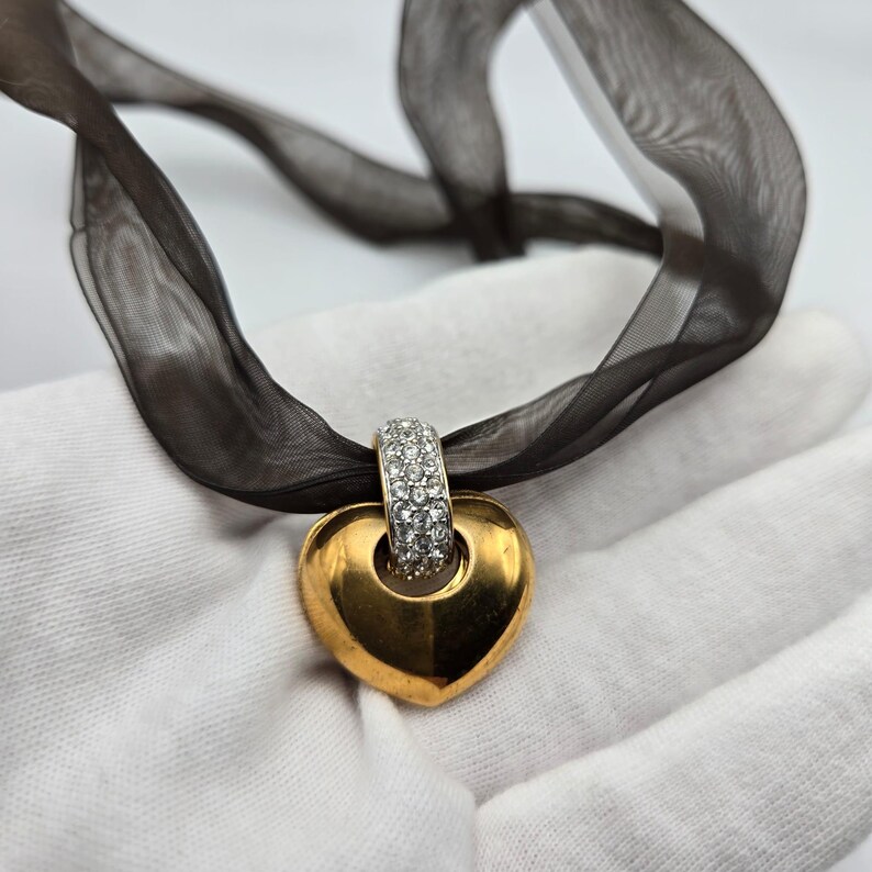 May include: A gold heart-shaped pendant with a clear crystal band around the top. The pendant is hanging from a black ribbon.