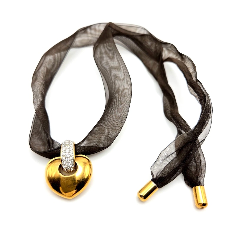 May include: A gold heart-shaped pendant with clear crystal accents hangs from a black ribbon necklace.