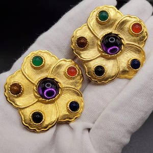 May include: A pair of gold tone flower-shaped earrings with five colorful gemstones in each earring. The center stone is a large purple stone, surrounded by four smaller stones in shades of green, red, brown, and blue.