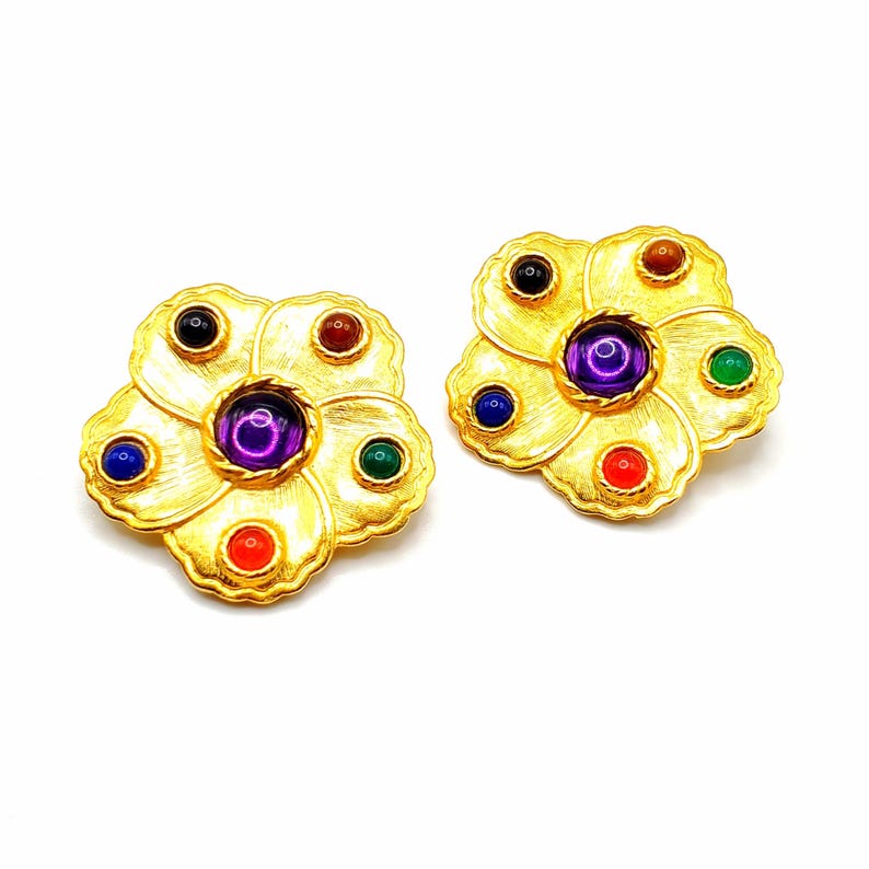 May include: A pair of gold tone flower-shaped earrings with multi-colored stones. The earrings have a central purple stone surrounded by five smaller stones in red, green, blue, and black.