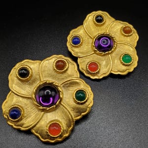 May include: A pair of gold tone flower-shaped earrings with multi-colored stones. The earrings have a central purple stone surrounded by smaller stones in red, green, blue, and black.