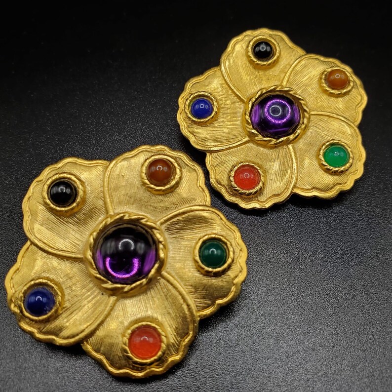 May include: A pair of gold tone flower-shaped earrings with multi-colored stones. The earrings have a central purple stone surrounded by smaller stones in red, green, blue, and black.