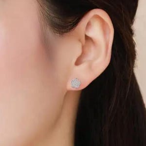 May include: A close-up of a person's ear with a small, silver flower-shaped stud earring. The earring is encrusted with small, sparkling stones. The skin is fair-toned, and the background is blurred.