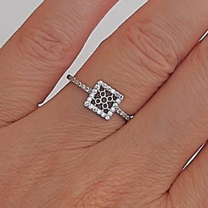 Può includere: Un anello d'argento con un design quadrato traforato. Il quadrato è delineato da piccole pietre chiare e il centro presenta un motivo di pietre più piccole e scure. La fascia è anch'essa ornata da piccole pietre chiare.