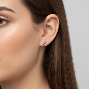 May include: A close-up of a woman's ear with a cluster stud earring. The earring features a small cluster of gemstones in red, blue, and green. The woman has long brown hair and fair skin.