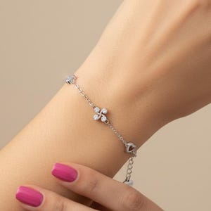 Pode incluir: Uma delicada pulseira de prata com um design floral com pedras preciosas transparentes. A pulseira tem uma corrente com um berloque de coração e uma pequena conta pendurada. A pulseira é exibida em um pulso com esmalte rosa.