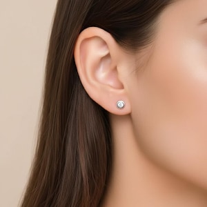 May include: A close-up of a woman's ear with a small, round, silver stud earring. The earring features a clear, sparkling gemstone set in a bezel setting. The woman has long, dark brown hair.