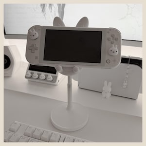May include: A white adjustable stand holding a handheld gaming console. The console features a white casing with small rabbit-shaped joystick covers. The stand has a circular base and a central support column, designed to hold the device securely.