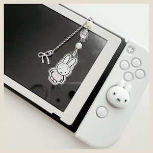 May include: A white gaming console controller featuring a white rabbit-shaped joystick cover. Attached to the console is a silver-toned chain charm with a clear acrylic rabbit pendant, a small silver bow charm, and white and clear spherical beads.