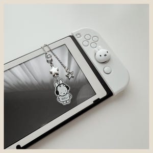 May include: A white gaming console controller featuring a small white rabbit-shaped thumbstick cover and a silver-toned metal chain charm. The charm includes a clear bead, a silver star, and a clear acrylic pendant with a black-lined rabbit illustration.