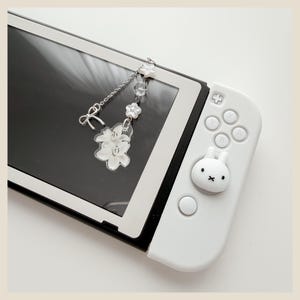 May include: A white gaming console controller featuring a white rabbit-shaped joystick cover and a silver-toned charm attached to the screen. The charm consists of a small bow, a silver chain, clear beads, and a white floral acrylic pendant.