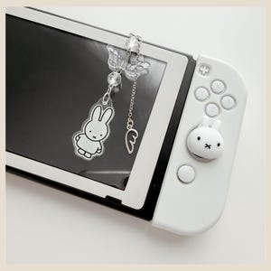 May include: A white handheld gaming console featuring a white rabbit character thumbstick cover and a dangling charm. The charm includes a clear acrylic rabbit pendant, a pair of clear wings, faceted beads, and a small silver wing charm on a chain.