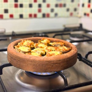 以下が含まれることがあります： ガスコンロの上に置かれた、焼いたキノコと溶けたチーズが入った丸いテラコッタの皿。チーズは黄金色。皿は点火されたバーナーの上にあります。背景にはタイルのバックボードがあります。