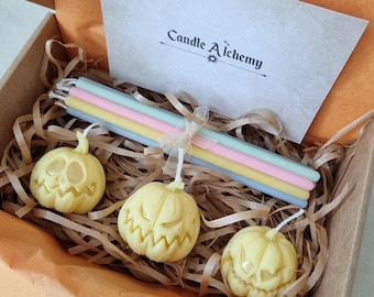 May include: A set of three small pumpkin-shaped candles in a box with shredded paper. The candles are yellow and have a carved jack-o'-lantern face. The box also includes a set of six colorful taper candles. The box has a card with the text "Candle Alchemy".