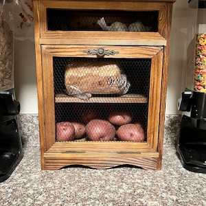 Könnte beinhalten: Ein Holzschrank mit Gitterfront, der Kartoffeln, Zwiebeln und Brot enthält. Der Schrank hat einen dekorativen Metallgriff und ist aus hellbraunem Holz gefertigt. Oben ist eine Packung englischer Muffins zu sehen.