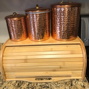 Op de afbeelding: Drie koperkleurige voorraadpotten met een gehamerde textuur en bijpassende deksels met goudkleurige handgrepen. De potten staan op een lichtgekleurde bamboe broodtrommel met een roldeksel. Keukenopslag.