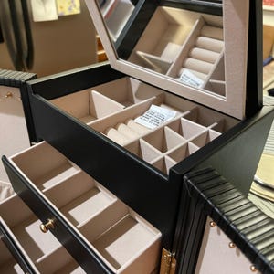 Pode incluir: Caixa de joias preta com vários compartimentos e gavetas. O interior é forrado com um tecido bege claro. A seção superior tem um espelho e suportes para anéis. Ferragens douradas acentuam a caixa.
