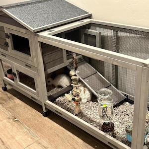 以下が含まれることがあります： グレーの屋根と金網の側面を備えた木製のウサギ小屋。小屋には複数のレベル、スロープ、水筒があります。内部は敷き材で満たされ、小さなウサギが中に見えます。