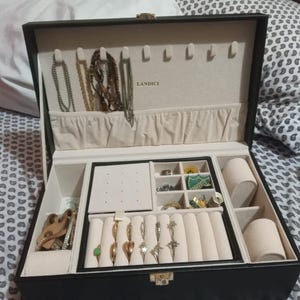 May include: Open black jewelry box with multiple compartments. The interior is lined with beige fabric and includes sections for necklaces, rings, and other jewelry. Various necklaces and rings are visible, along with the brand name "LANDICI".