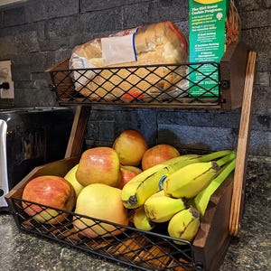 May include: A two-tiered wooden and metal wire basket filled with food. The top level holds a loaf of bread and a box of Girl Scout Cookies. The bottom level contains apples and bananas. The basket is on a countertop.