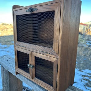 Könnte beinhalten: Kleiner Holzschrank mit natürlicher Oberfläche. Der Schrank hat eine Gitterfront im oberen Bereich und zwei Türen unten. Dekorative Metallbeschläge sind sichtbar. Der Schrank steht auf einem Holzzaun.