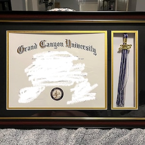 Puede incluir: Diploma enmarcado de la Universidad Grand Canyon, con una borla dorada del año 2023. El marco es negro con un borde interior dorado y un borde exterior marrón oscuro. El diploma es parcialmente visible.