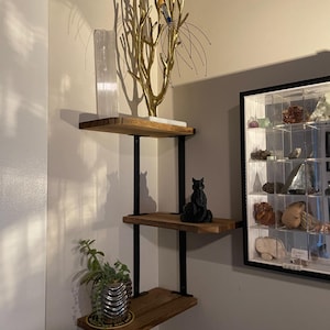 Peut inclure: Étagère d'angle à trois niveaux avec étagères en bois et supports en métal noir. L'étagère supérieure contient un arbre décoratif doré et un cristal blanc. L'étagère du milieu affiche une figurine de chat noir. L'étagère inférieure a une plante en pot.