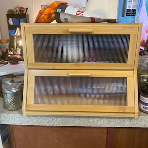 Op de afbeelding: Een bamboe brooddoos met twee niveaus en heldere, geribbelde glazen deuren. De doos heeft een natuurlijke houten afwerking en rechthoekige handgrepen. Bovenop liggen een zak chips en een container.