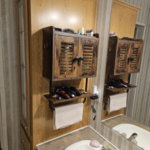 Op de afbeelding: Een houten badkamerkast met een verweerde afwerking, met een kast met twee deuren, een plank en een handdoekenrek. De kast is gemonteerd op een licht houten wandpaneel boven een wastafel. Diverse verzorgingsproducten staan op de plank.